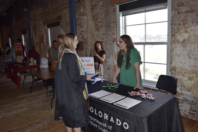 A woman stands at a Colorado Department of Higher Education branded table and smiles at a young woman wearing a cap and gown.