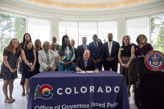 Group of officials and staff gathered around a table with a Colorado state emblem, where a man is signing a document in a bright room.