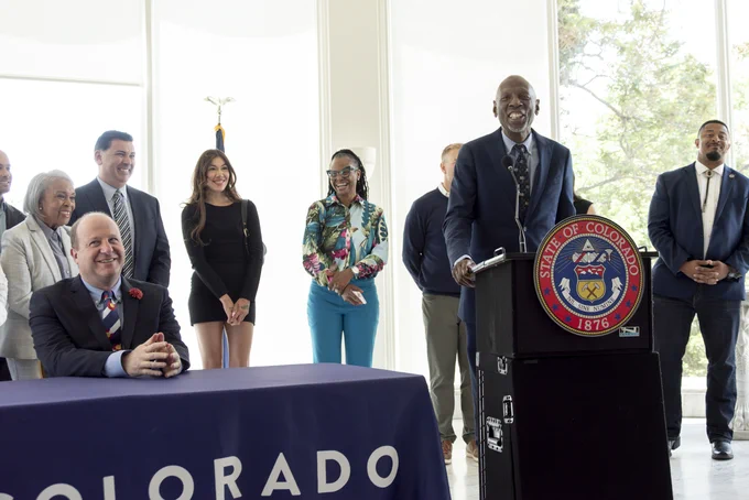 A man in a suit speaking at a podium with the Colorado state seal, surrounded by a diverse group of people standing behind a table covered with a Colorado banner.
