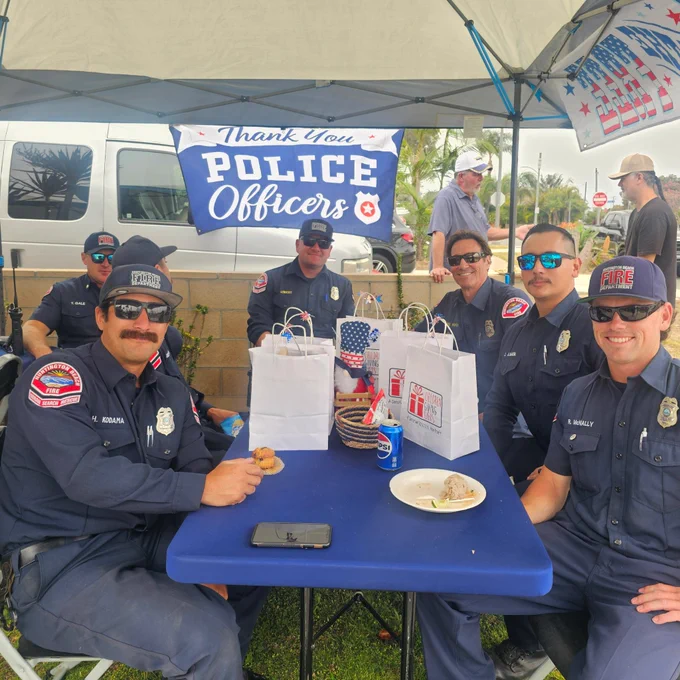 Huntington Beach firefighters gather under a canopy during a community appreciation event. A banner thanking police officers hangs in the background as firefighters visit, share a meal, and receive gift bags from community members.