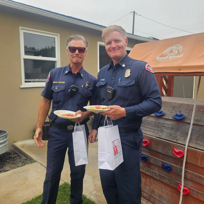 Huntington Beach Fire Captains Luke Jones and Berkley Northrop stand outdoors holding meals and gift bags during a community appreciation event. The captains smile for a photo while visiting with community members and supporters who gathered to recognize local first responders.
