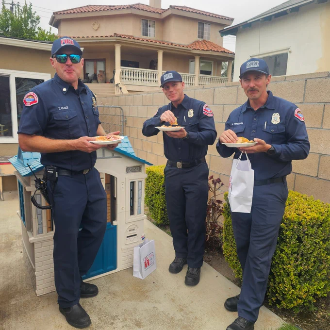 Huntington Beach firefighters Joe Ferguson, Jordan Lasher, and Tristan Gale enjoy a meal during a community appreciation event. The firefighters stand together outdoors while visiting with community members and showing appreciation for the support provided to local first responders.
