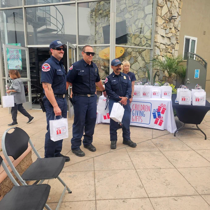 Huntington Beach firefighters Donny Sand, Ronny Perez, and Kevin Martin pose for a photo during a community appreciation event hosted by Children Giving Gifts. The firefighters hold gift bags while standing near a display table prepared by volunteers who organized the event in support of local first responders.