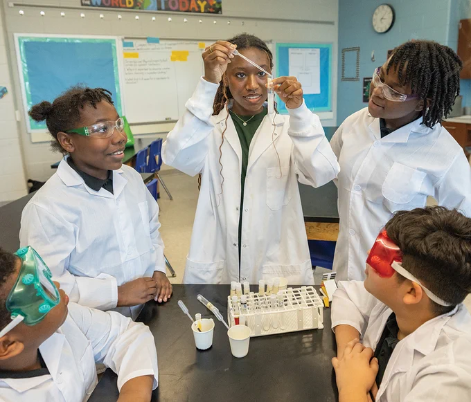 A group of students in a classroom wearing safety goggles and lab coats are gathered around a table. One student is holding up a dropper, while others observe. Test tubes and a rack are visible on the table. The room has educational posters and a clock on the wall.