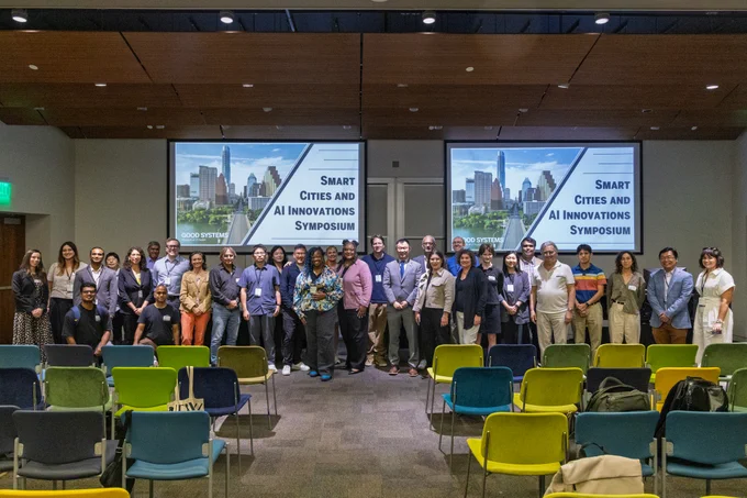 Group photo of attendees and speakers at the Smart Cities and AI Innovations Symposium in a UT Austin auditorium. Two screens behind the group display the symposium title graphic, and rows of colorful chairs are visible in the foreground.