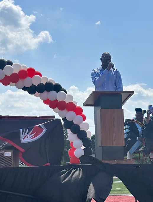 Lieutenant Governor Garlin Gilchrist II stands at a wooden podium speaking into a microphone during an outdoor graduation ceremony. He is wearing a light blue button-down shirt. To his left, a large decorative arch made of red, white, and black balloons curves over the stage, set against a bright, partly cloudy blue sky.