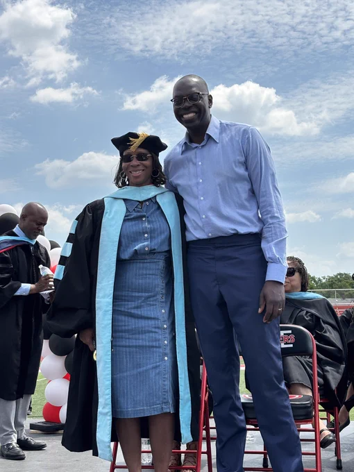 Lieutenant Governor Garlin Gilchrist II, wearing a light blue button-down shirt and navy trousers, smiles warmly with his arm around a woman wearing a denim dress and a black and light blue academic graduation gown and cap. They are standing outdoors on a stage under a partly cloudy sky, with other graduates and a red, white, and black balloon arch visible in the background.