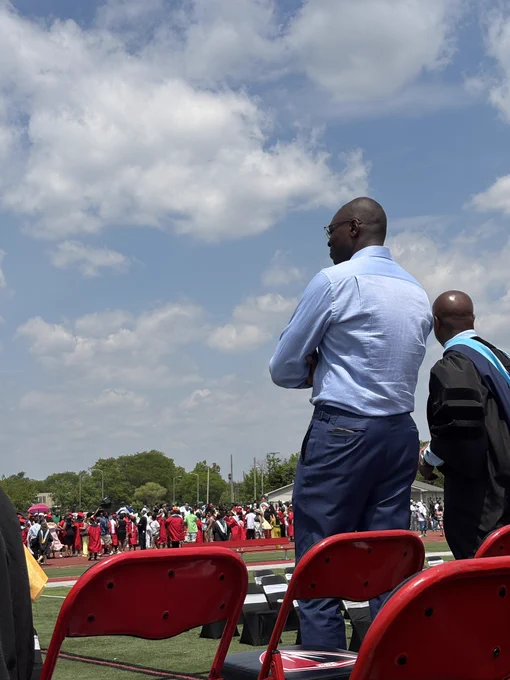 A rear side view of Michigan Lieutenant Governor Garlin Gilchrist II, dressed in a light blue shirt and dark trousers, standing with his arms crossed on an outdoor stage. He is looking out at a large crowd of graduates dressed in red and black gowns gathered on a sports field. Red folding chairs are visible in the immediate foreground under a blue sky with scattered clouds.