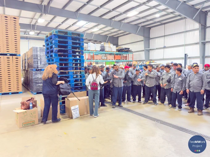 Workers in uniform stand together inside a produce facility while TeaMWork Project staff hand out backpacks from teamwork kits during an outreach visit.
