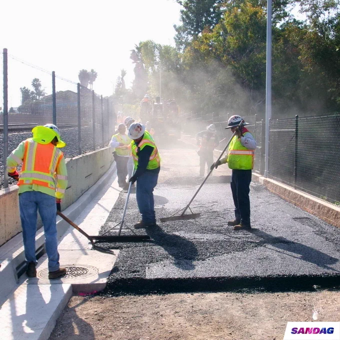Workers in reflective vests and helmets are paving the Inland Rail Trail. The SANDAG logo is visible in the bottom corner.