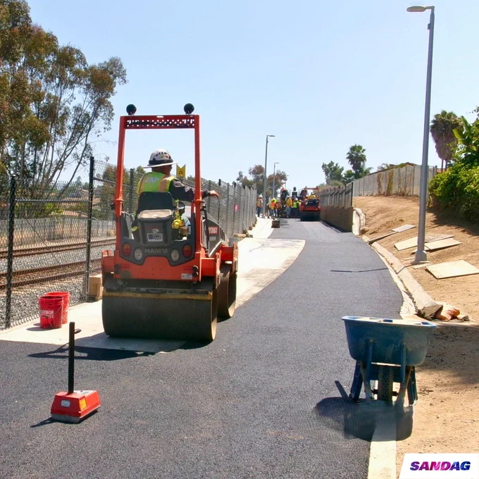 A worker operates a steamroller on the Inland Rail Trail path. Additional workers and machinery are visible further down the path. The SANDAG logo is present in the lower corner.