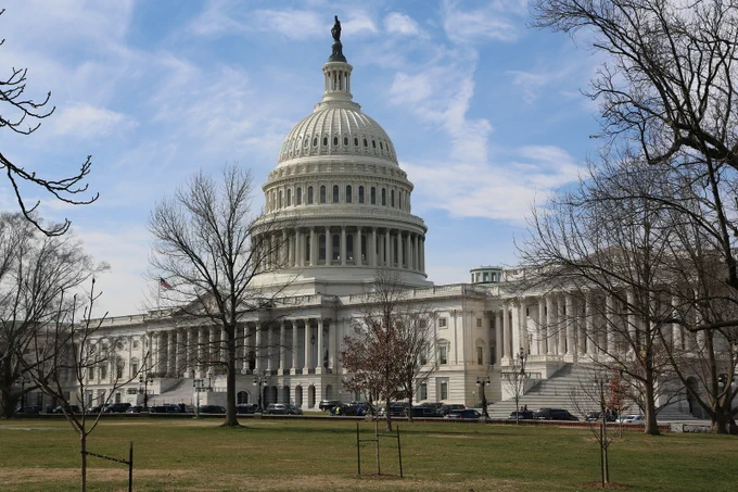 A view of the US Capitol Building surrounded by bare trees under a blue sky. The mood is solemn and historic.