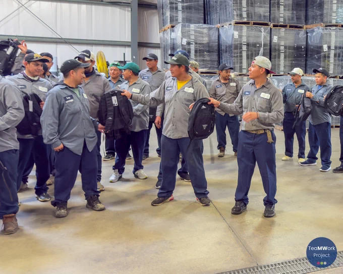 Large group photo inside a warehouse with migrant agricultural workers holding backpacks and a Guatemalan flag alongside outreach partners during the Windsor-Essex visit.
