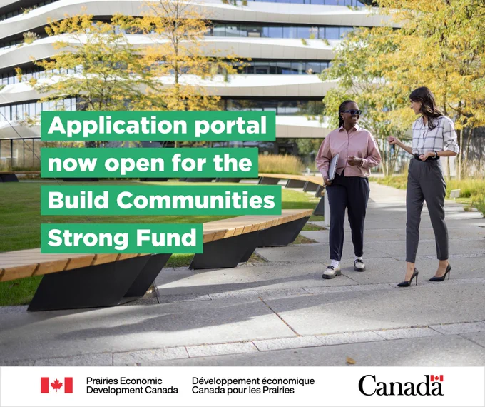 Two business women are walking through a park talking to each other with business buildings in the background. One woman holds a laptop. With text: “Application portal now open for the Build Communities Strong Fund”. 