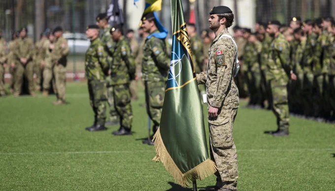 Canadian soldier in uniform holding a NATO flag during a military ceremony on a grassy field.