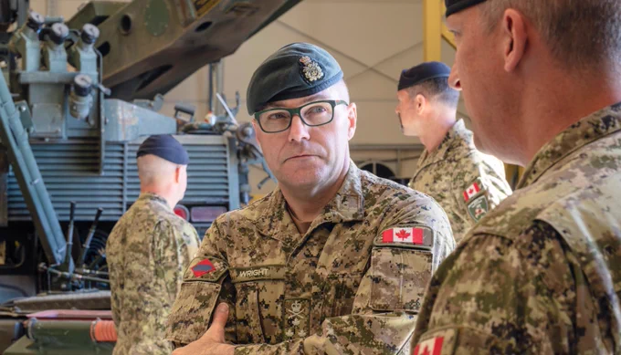 Canadian military personnel in camouflage uniforms and berets engaged in conversation inside a vehicle maintenance facility.