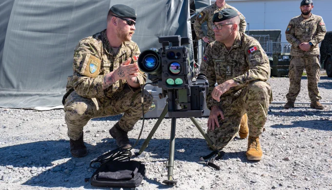 Two soldiers in camouflage uniforms operate a tripod-mounted advanced optical targeting device outdoors on gravel ground. 