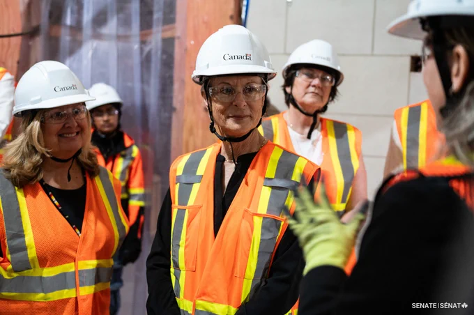 A group of people wearing safety vests and helmets, construction works visible in the background.