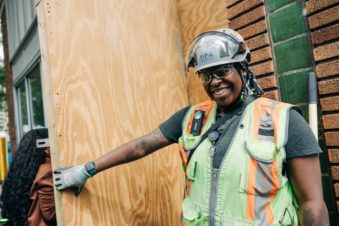 A woman in a hard hat holds a plywood door as those from the tour enter the building. 