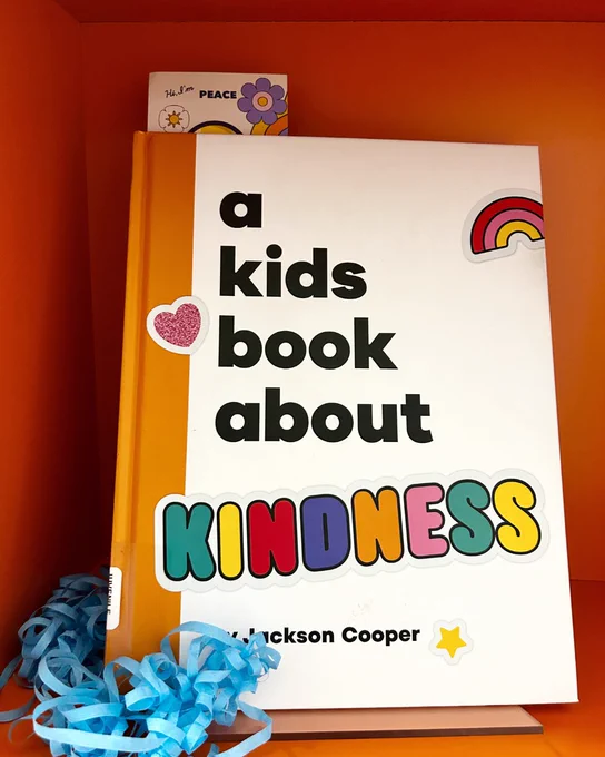 A book titled "a kids book about kindness" displayed on an orange shelf with a bookmark peeking out