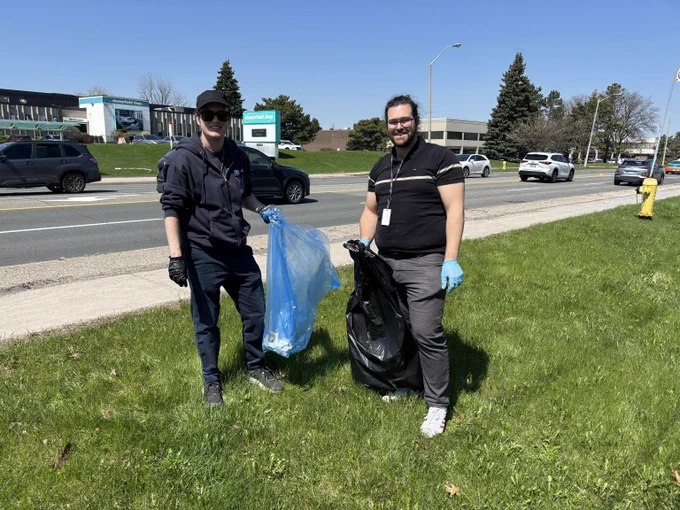 Two people holding trash and recycling bags. A street with cars and shops in the background.