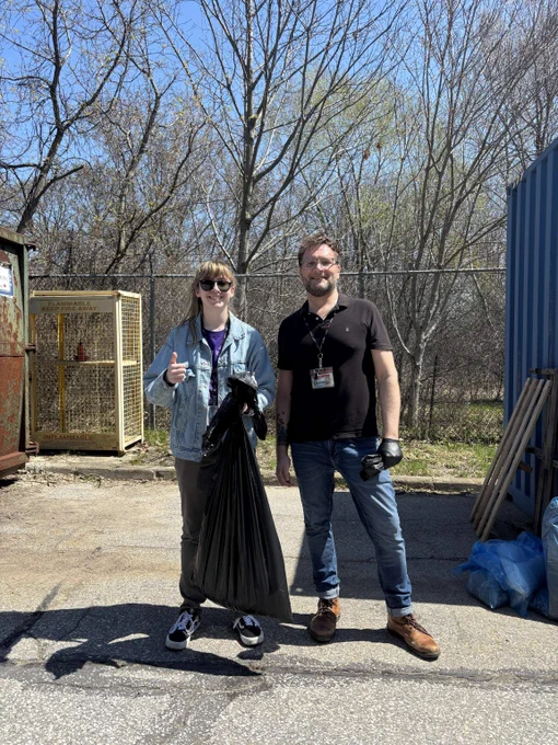 Two people holding trash bags, surrounded by trash and trash containers. A fence and trees behind them.