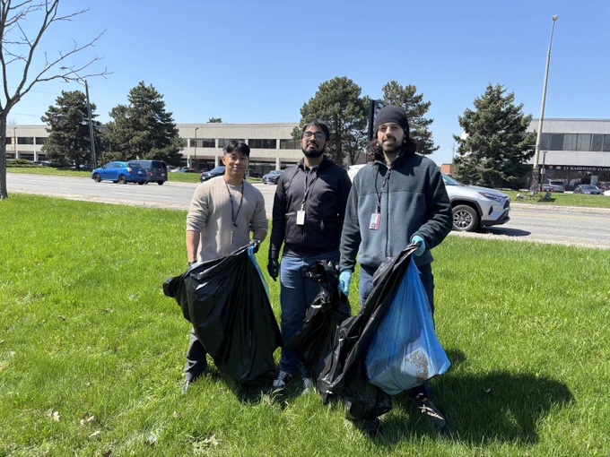 Three people holding trash and recycling bags. A street with cars passing by and buildings in the background. 