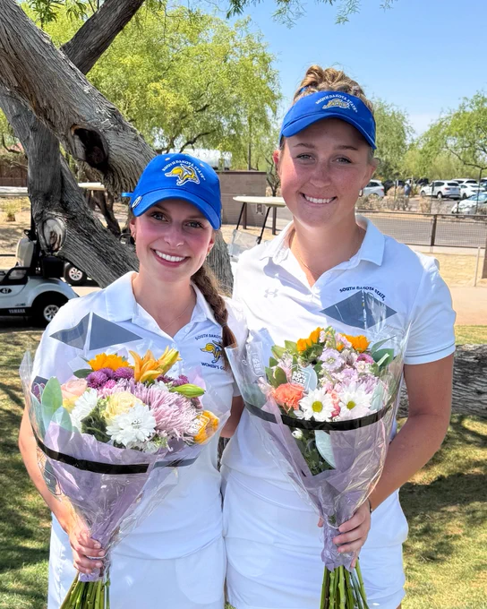 Two women’s golfers stand side-by-side, wearing matching white golf outfits and blue hats, holding flower bouquets