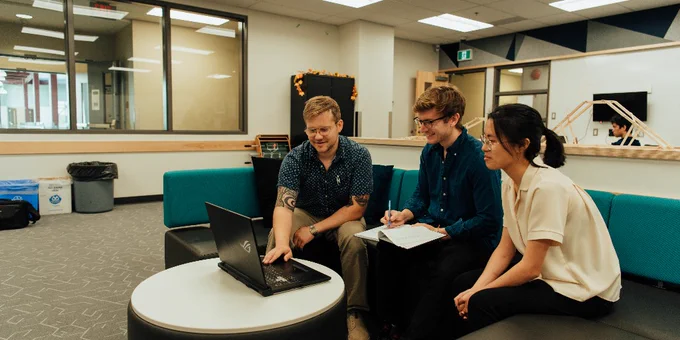 A group of three individuals sits on turquoise couches in an office setting, engaged with a laptop on a small round table. The room has large windows, gray carpet, and neutral-colored walls.