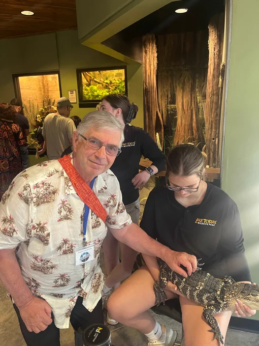 Man in a floral shirt and red scarf touching a small alligator held by a seated person in a black shirt inside an indoor exhibit area.