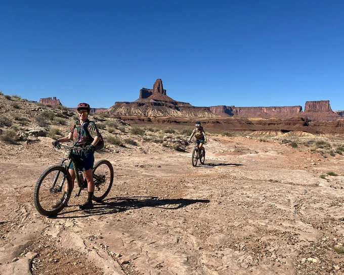 Two adults biking through a desert landscape with a large rock formation in the far background.