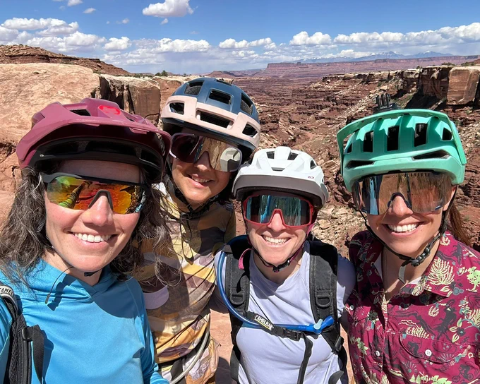 Four adults in bike helmets and sunglasses smile at the camera while taking a selfie.
