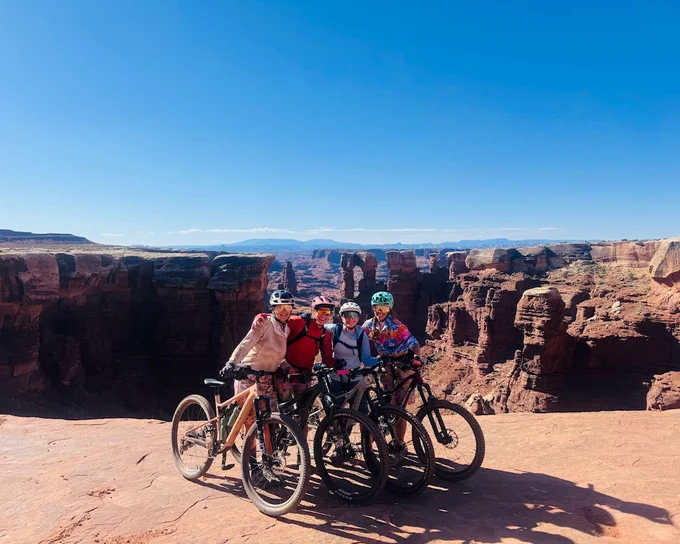 Four adults in biking gear and mountain bikes pose together at the edge of a canyon.