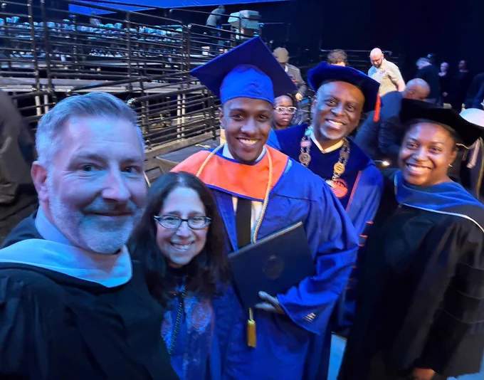 A group photo taken at a graduation ceremony. Five people are smiling, with one dressed in a blue cap and gown, holding a diploma. Another person is wearing a ceremonial medallion over their gown. The setting includes seating in the background, indicating an event space.