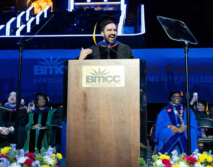 NYC Mayor Zohran Mamdani in graduation attire stands at a podium with the BMCC logo, speaking at a commencement ceremony. Behind, a group of people in academic regalia are seated. A large screen displaying colorful lights is in the background. Bright flowers decorate the front of the stage.