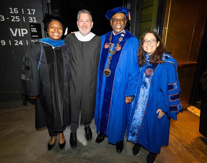Four people in academic regalia stand closely together, smiling for a photo. Two individuals are wearing dark gowns and caps, while the other two are in bright blue robes with ornate designs. One person is wearing an academic chain of office. They are posed in front of a black wall with VIP signage.