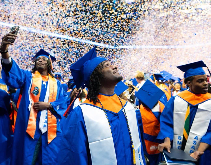 Graduates in blue and orange gowns and caps celebrate at a commencement ceremony, with a flurry of confetti filling the air. People are taking photos and looking upward.