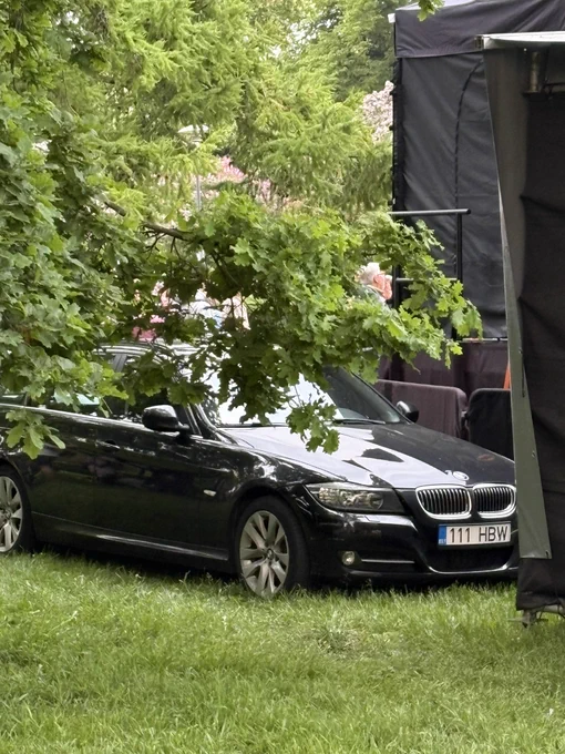 Fotol on must BMW universaalkerega sõiduauto, mis on pargitud murule suure puu alla. Auto on osaliselt varjatud madalale vajunud tammeoksa poolt, mille lehed katavad osa katusest ja esiklaasist. Paremal paistab musta kattega ajutine lava või ürituse konstruktsioon. Taustal on rohelised puud ning aimatavalt inimesed. Pilt on tehtud pargis või üritusalal ning jätab mulje, nagu oleks auto pargitud otse haljasalale puude vahele.