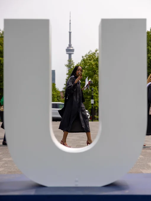 UTSC graduate walks through a large white letter "U" sculpture in convocation. In the background, the CN Tower is visible, surrounded by greenery. 