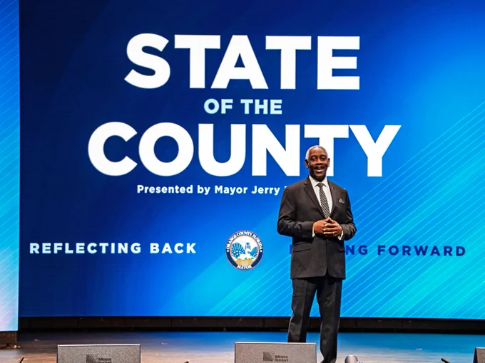 The image shows Orange County Mayor Jerry Demings on stage in front of a large screen displaying the text "State of the County, Presented by Mayor Jerry Demings"