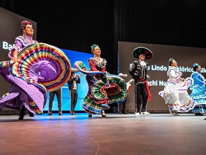 Performers in vibrant, traditional Mexican attire dance on stage at the Orange County State of the County event. Swirling skirts create colorful patterns, and one dancer wears a sombrero. 
