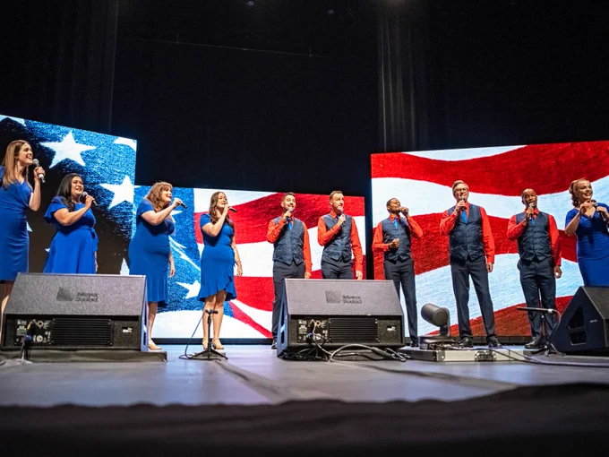 The Epcot Voices of Liberty group, dressed in blue, singing into microphones at the Orange County State of the County event. Behind them are large screens displaying the American flag.