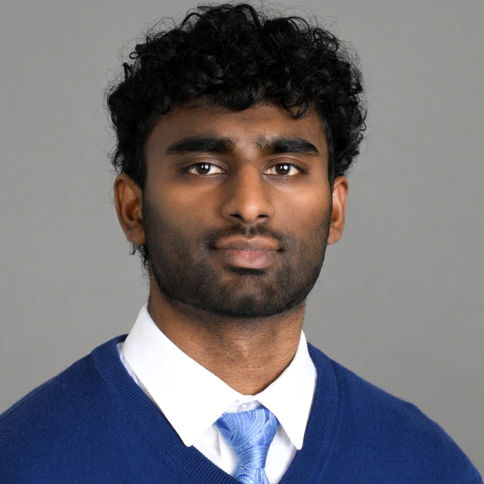 Joshua Mathew wearing a blue sweater and tie with a white collared shirt in front of a gray background