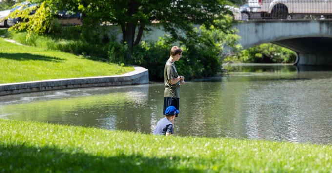 A person wearing a green shirt is standing while another is crouching in the grass on the human-made shore of a lake as full-leaf trees and other foliage nearly obscure a bridge in the background. 