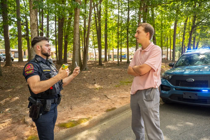 A police officer is standing and talking to an individual near a Ford police vehicle with emergency lights on, in a wooded area. The officer is holding a breathalyzer and speaking to the person, who is standing with arms crossed.