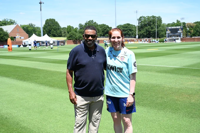 UNCG Chancellor Franklin D. Gilliam, Jr., and Norwegian Football Federation President Lise Klaveness stand on a sunny soccer field with green grass and a scoreboard in the background.