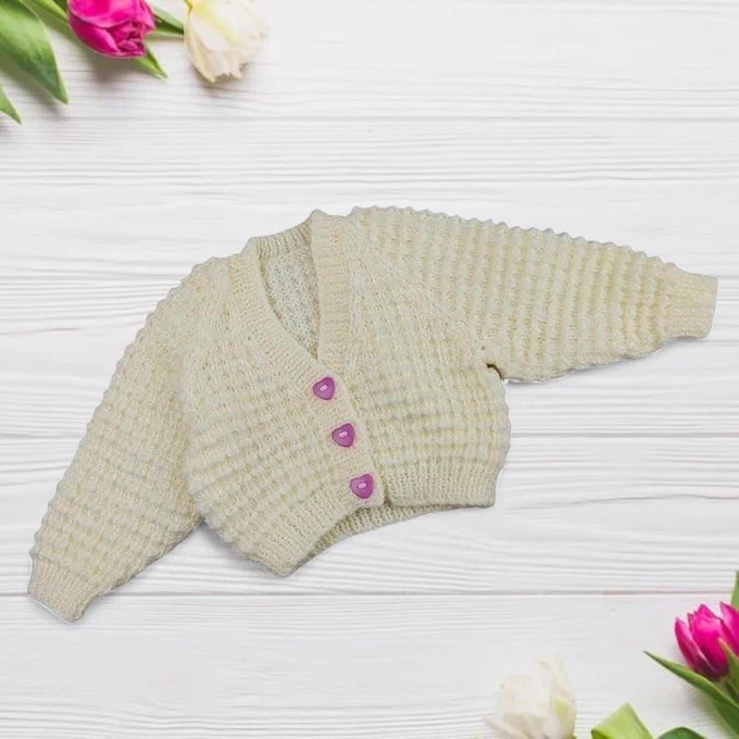 A cream knitted baby cardigan with three pink heart buttons lies centered on a white wooden plank background. The texture is a bumpy waffle stitch. Vibrant pink and white tulips are arranged in the top left and bottom right corners.