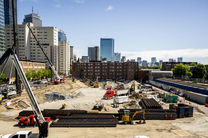 A bustling urban construction site under a clear blue sky. Dirt mounds, excavators, large cranes, and stacks of metal pipes fill the foreground. A KELLER piling rig is visible on the left. Tall skyscrapers and older brick buildings create a dense city skyline in the background. Workers are near a red truck.