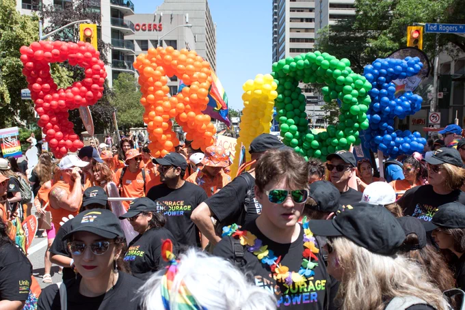 Crowded street parade scene with large balloon letters spelling “PRIDE” in red, orange, yellow, green, and blue above a group of participants.