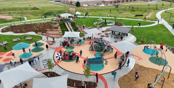An aerial view of a vibrant playground filled with various play structures and equipment. People are scattered around the area, engaging in different activities. There are multiple slides, climbing nets, and swings. The playground is surrounded by green spaces and winding paths, and some people are sitting under large shade umbrellas.